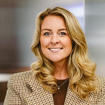 Woman with long blonde hair wearing a brown checked blazer and dark top, smiling at the camera in an indoor setting.