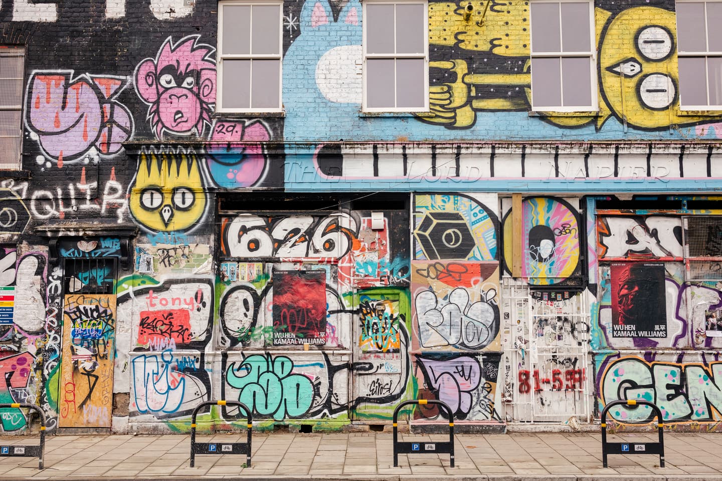 A building façade covered in colourful graffiti art, tags, and posters, with boarded-up windows and doors, and two bicycle racks on the pavement in front.
