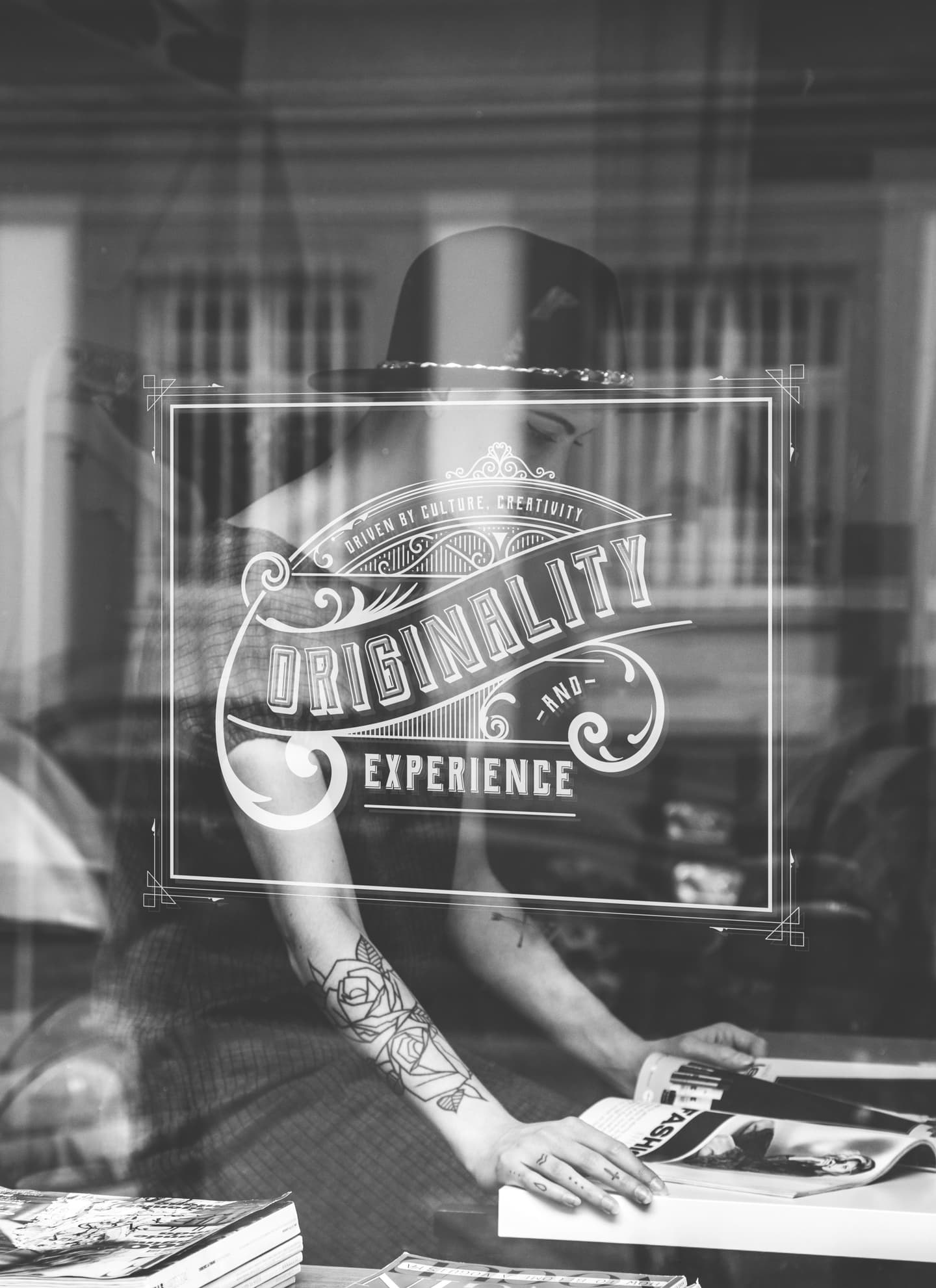 Person with tattooed arm sits at a table with open books behind a glass window featuring the words Originality and Experience.
