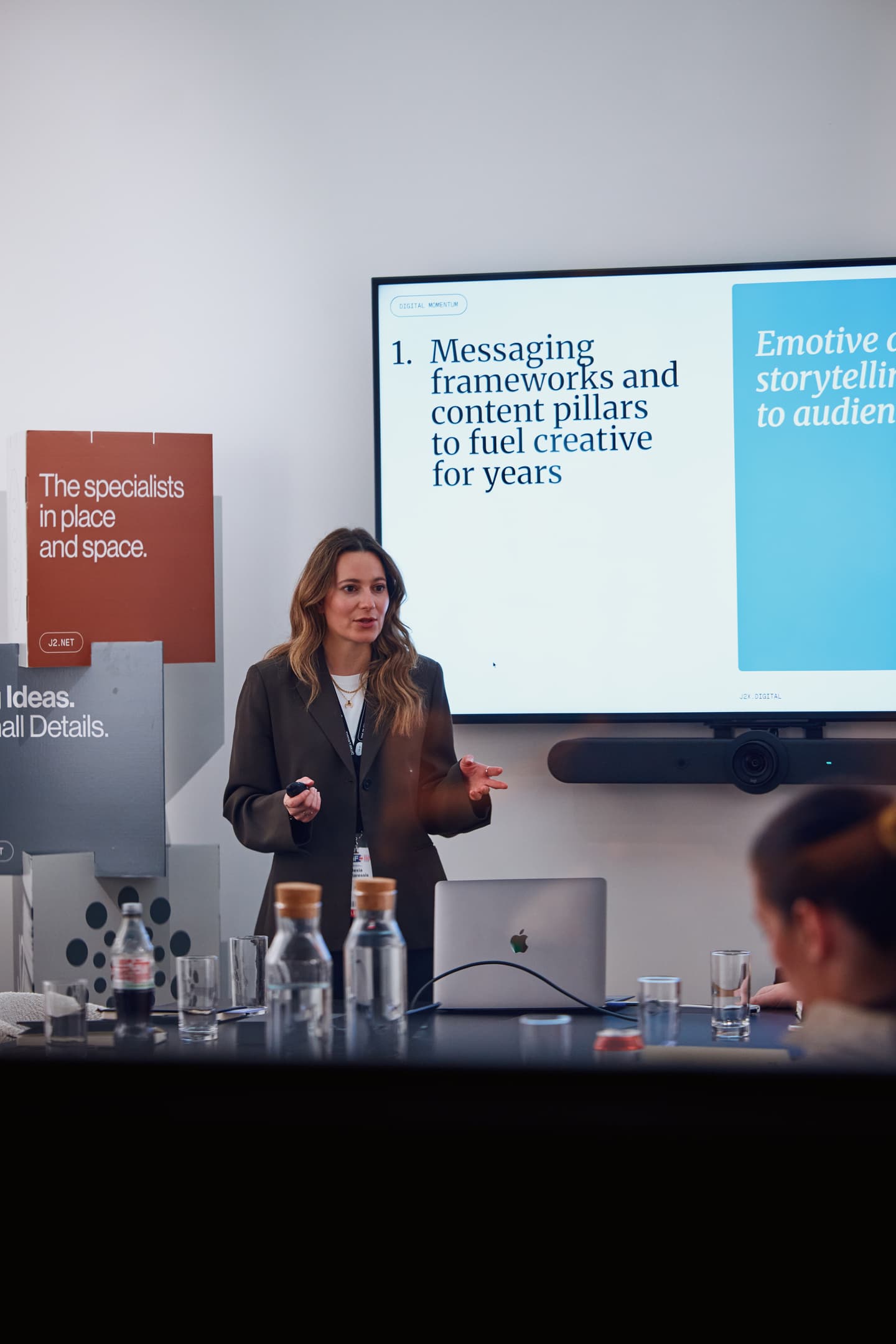 A woman gives a presentation in a conference room, standing in front of a screen displaying a slide about messaging frameworks and content pillars.