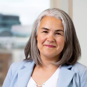 A woman with shoulder-length grey hair, wearing a light blue blazer and white blouse, stands indoors in front of a window with an urban background.