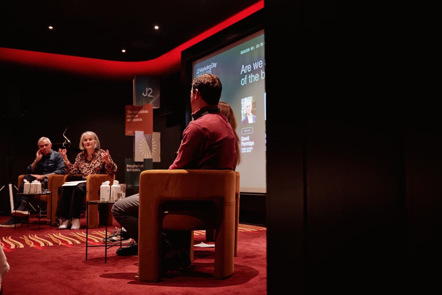 Four people participate in a panel discussion in a dimly lit room with a projected presentation behind them and audience members out of shot.