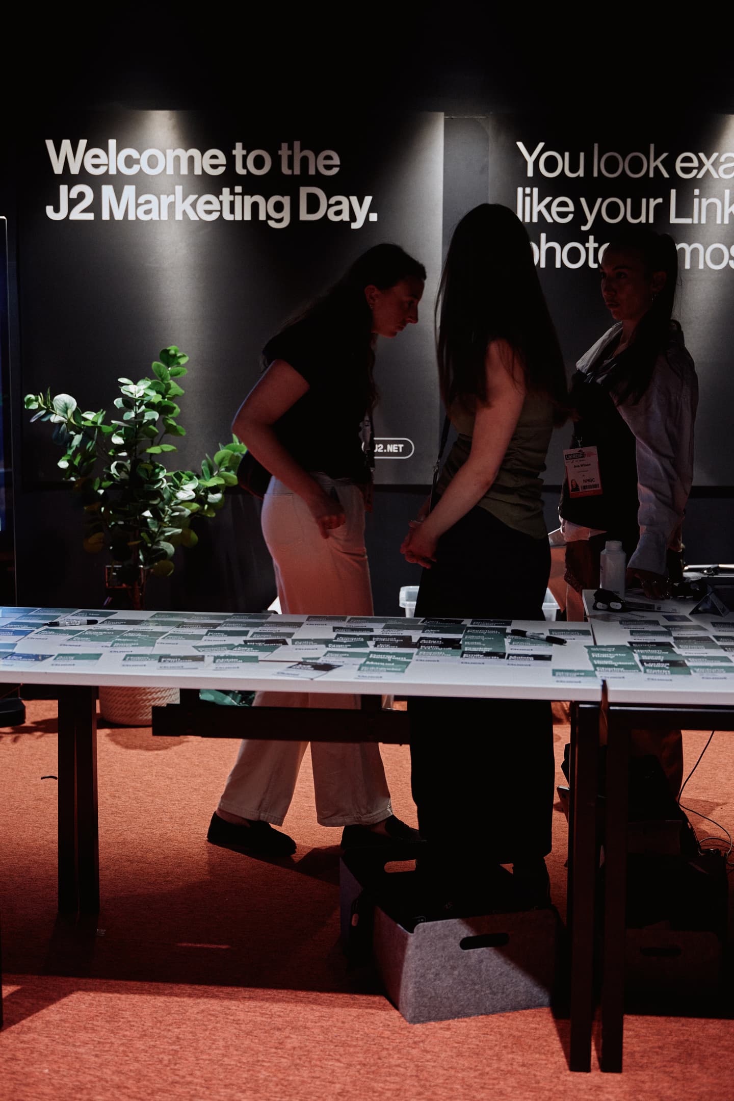 Three people stand near a registration table covered with name badges at an event with a sign reading Welcome to the J2 Marketing Day on the wall behind them.