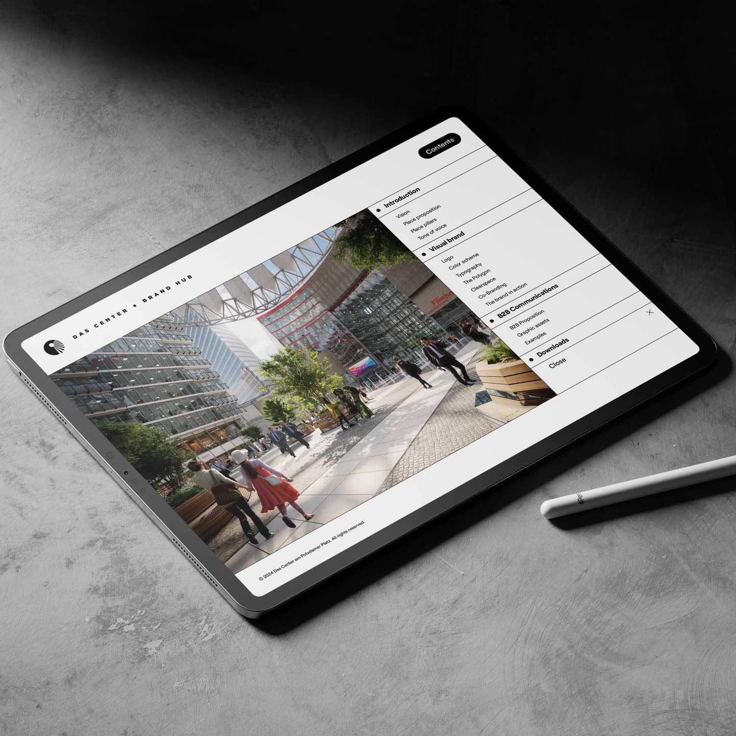 A tablet on a grey surface displays an architectural rendering of a modern outdoor plaza with people walking and interacting.