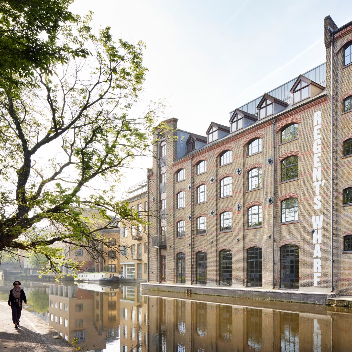 A person walks along a canal beside a brick building labelled Regents Wharf, with trees and moored boats visible.