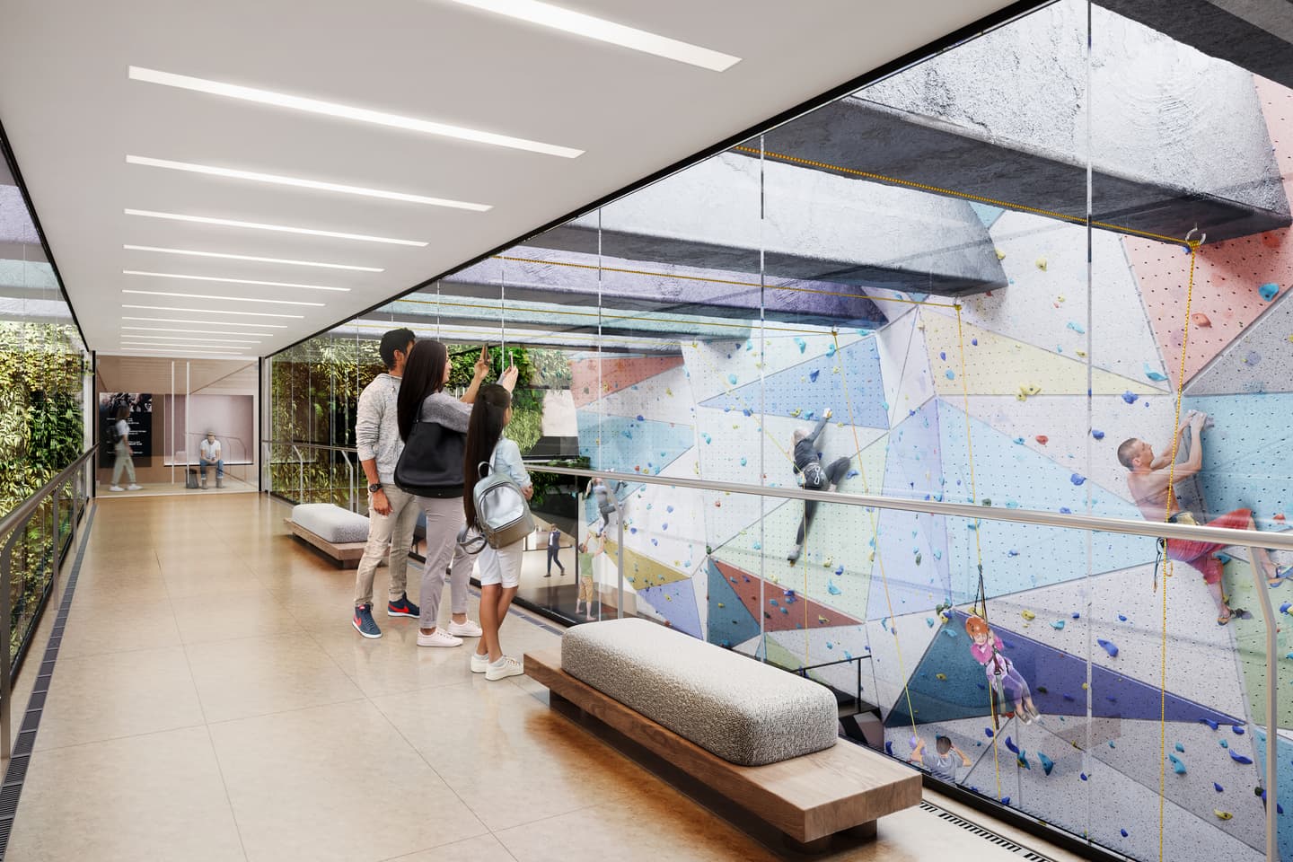 People stand in a glass corridor observing children and adults climbing a colourful indoor rock climbing wall on the other side of the glass.