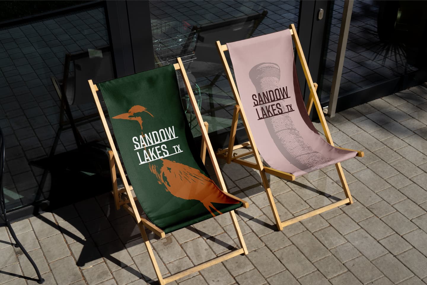 Two wooden deck chairs with fabric seats are positioned on a patio. The green chair features a bird, and the pink chair displays an oil well and both read SANDOW LAKES TX..