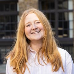 A person with long red hair, wearing a light-coloured button-up shirt, smiles whilst standing outdoors in front of a building with large windows.