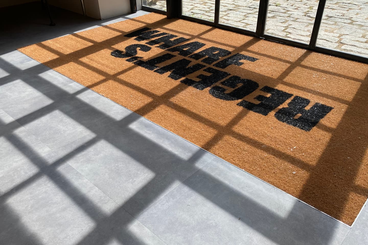 A brown doormat with REGENTS WHARF printed in black, seen upside down, lies near glass doors. Sunlight casts window grid shadows on the mat and grey floor.