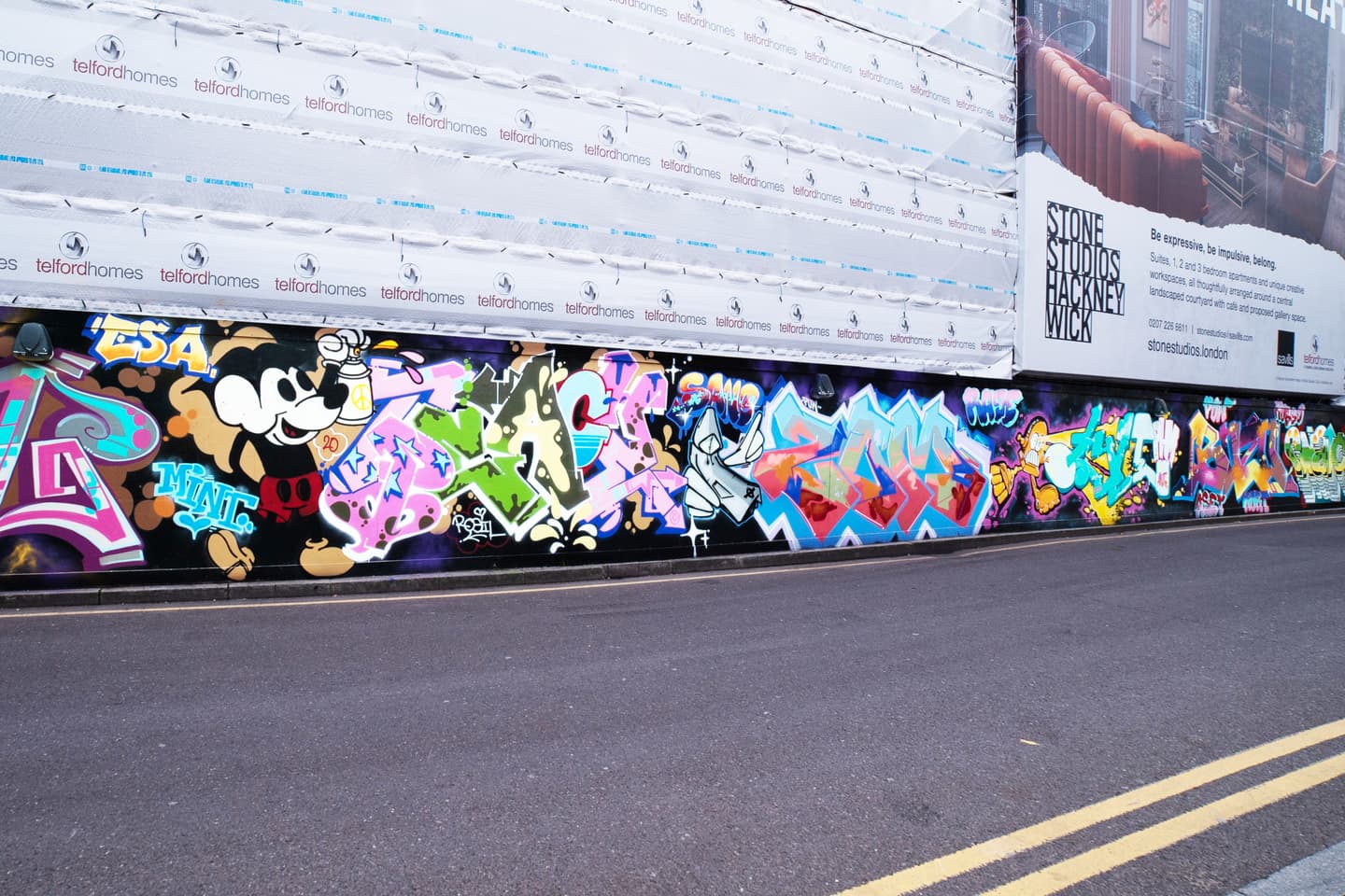 A long mural featuring colourful graffiti art, including a cartoon character, covers a construction site hoarding along a city street with double yellow lines.