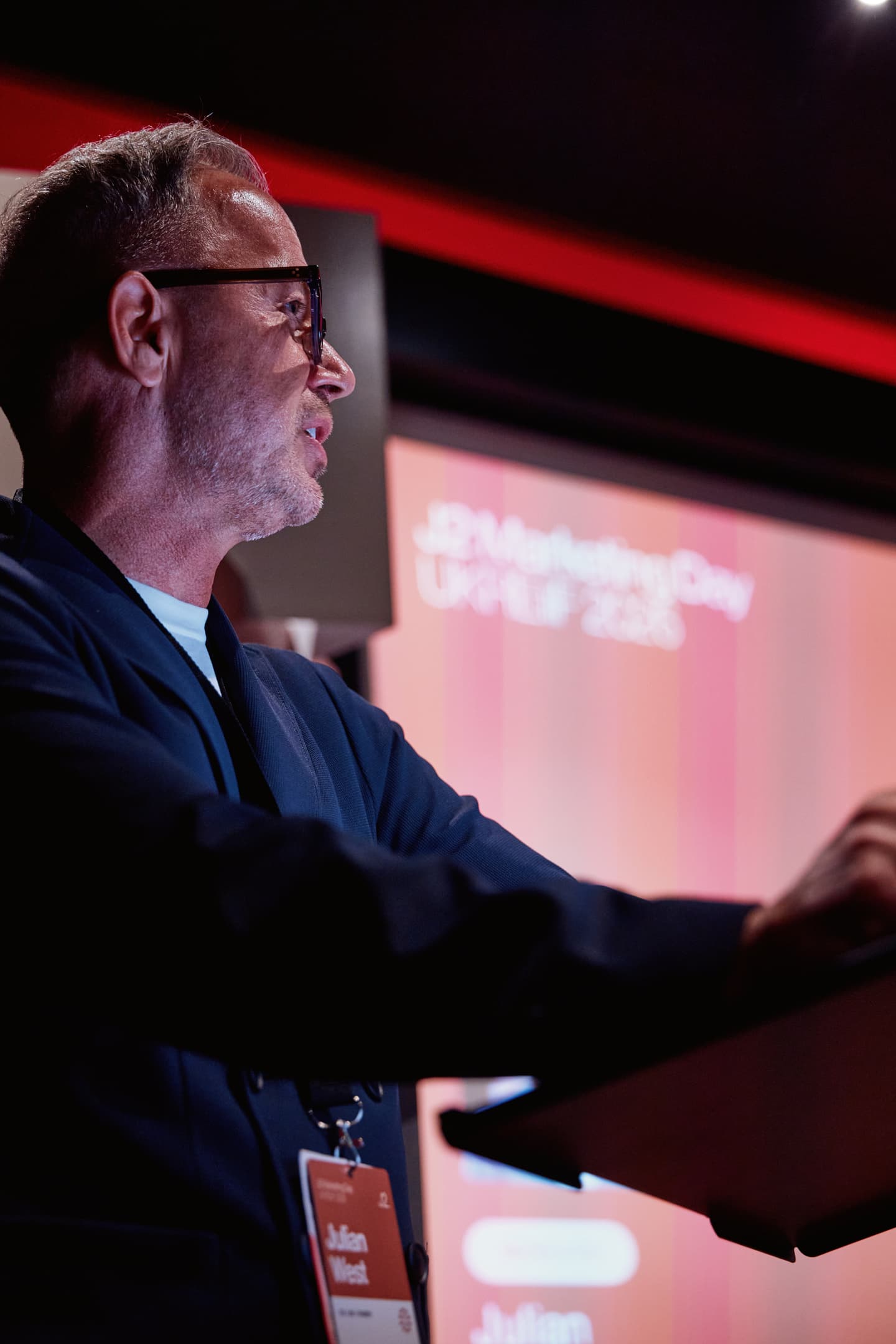 A man wearing glasses speaks at a lectern during a marketing event, with a blurred presentation screen in the background.