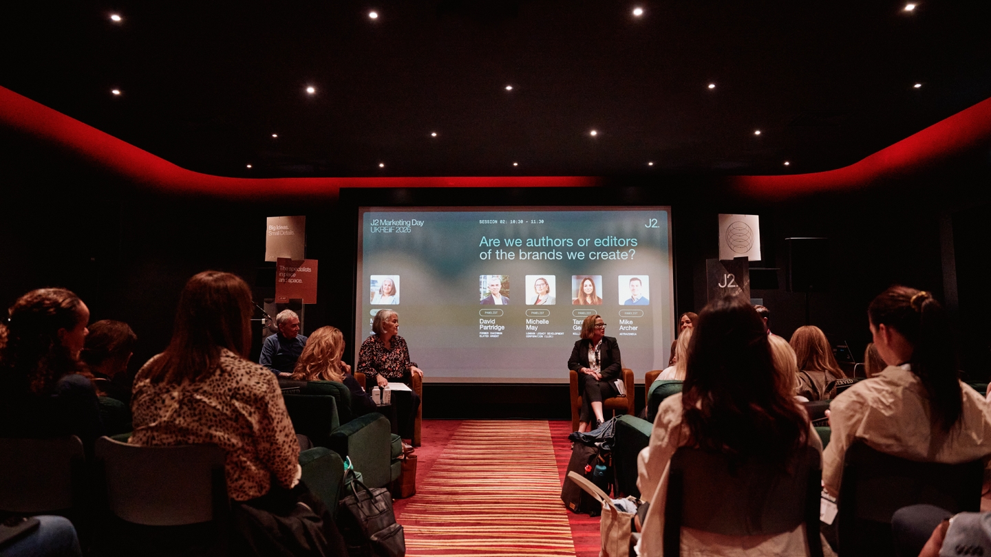People sit in a dimly lit auditorium watching a panel discussion with four speakers onstage; a large screen behind them displays topic details and speaker names.