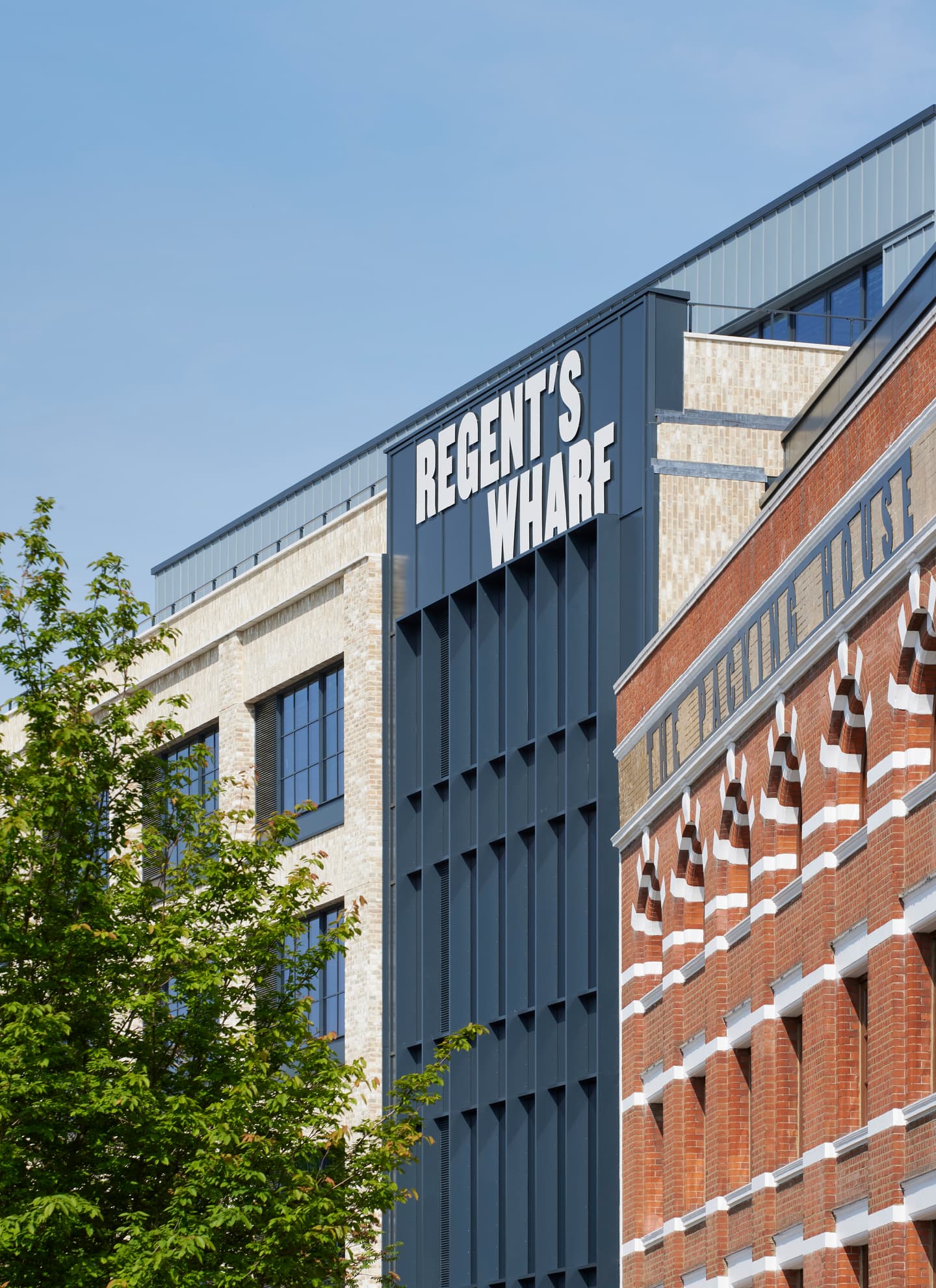 Modern Regents Wharf building with dark panels and large windows next to a red-brick building, tree in foreground, clear sky above.