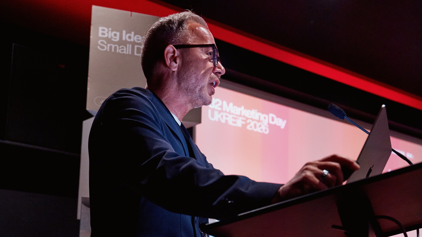 A man in a suit speaks at a lectern with a microphone during a marketing event; event signage and red lighting are visible in the background.