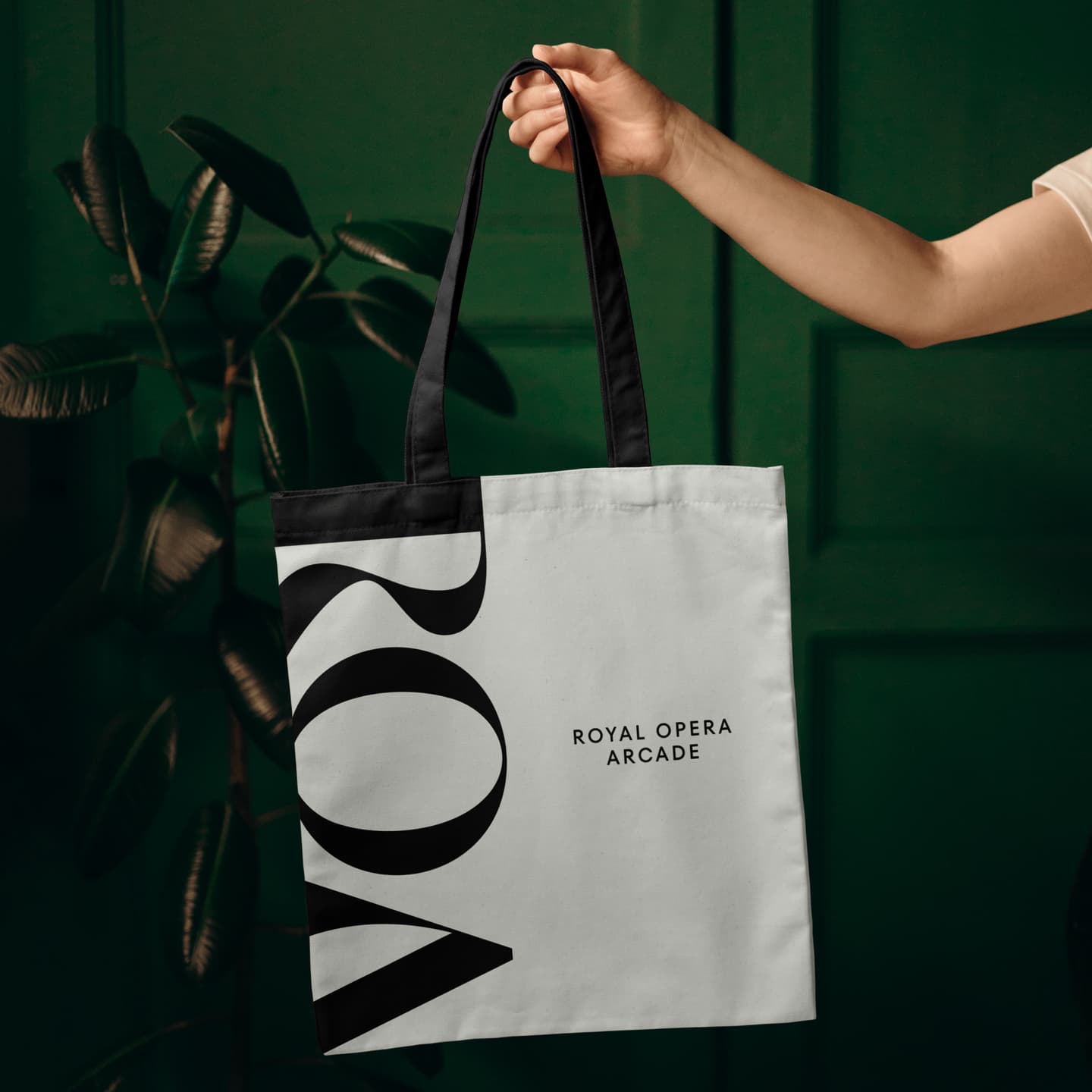 A hand holds a white and black tote bag with ROYAL OPERA ARCADE and large partial letters ROA printed on it, with a green plant and wall in the background.