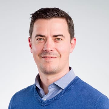 A man with short dark hair wearing a blue jumper over a collared shirt, smiling slightly, posed against a plain light background.