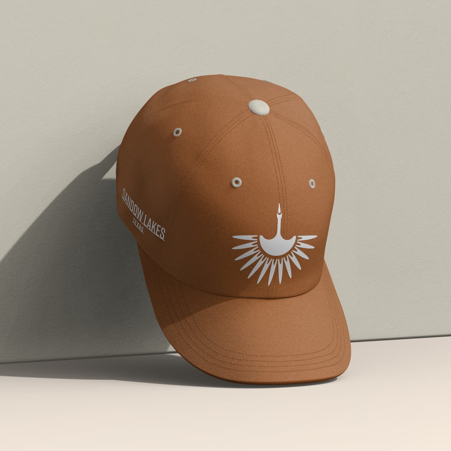 Brown baseball cap with a white abstract sunburst design on the front and the text SANDON LAKES RESORT on the side, resting against a light-coloured wall.