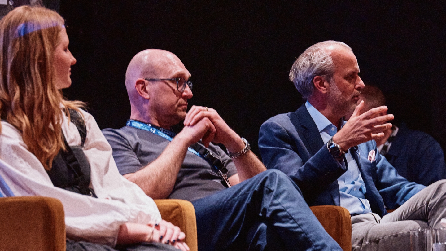 Three people sit on stage in a panel discussion. The man on the right gestures whilst speaking. The other two listen attentively. The background is dark.