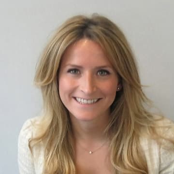 A woman with long, blonde hair is smiling at the camera. She is wearing a light-coloured top and a small necklace, with a neutral background behind her.