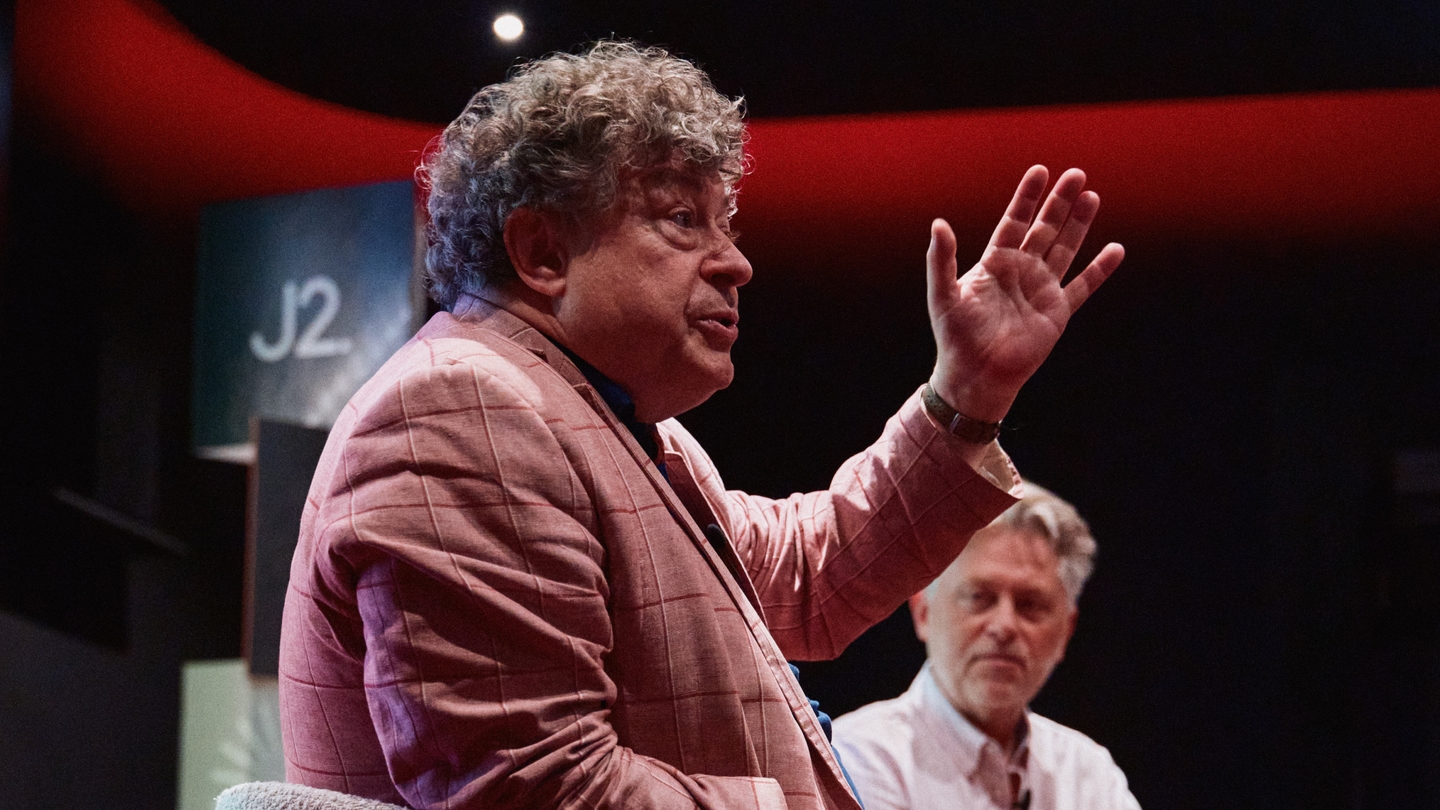 A man in a pink checked jacket gestures with his hand while speaking, as another man in a white shirt sits beside him in a dimly lit room.