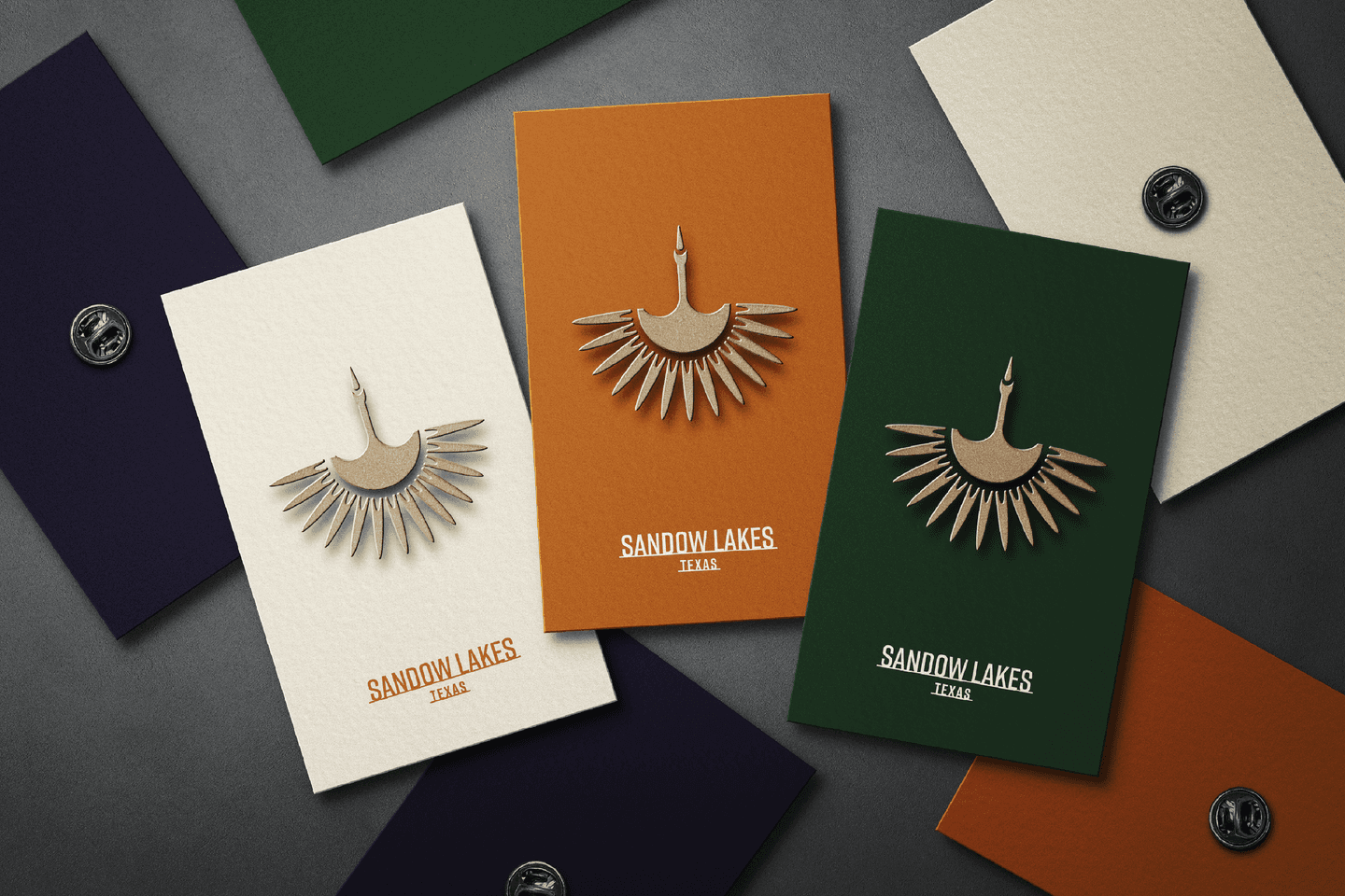 Four cards with bird-shaped pins and SANDOW LAKES TEXAS text are displayed on a dark surface, each card in different colours: navy, orange, white, and green.