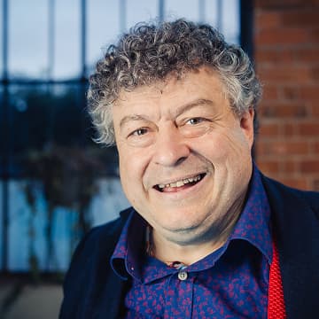 A middle-aged man with curly grey hair smiles at the camera, wearing a blue patterned shirt and blazer, standing indoors near a window with a brick wall in the background.