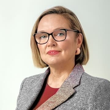 A woman with short blonde hair, glasses, and a grey blazer over a red top poses against a plain light background.