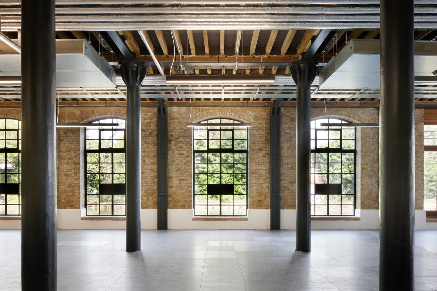 Interior of an industrial-style room with exposed brick walls, metal beams, large windows, and ceiling ducts. Natural light fills the empty space.