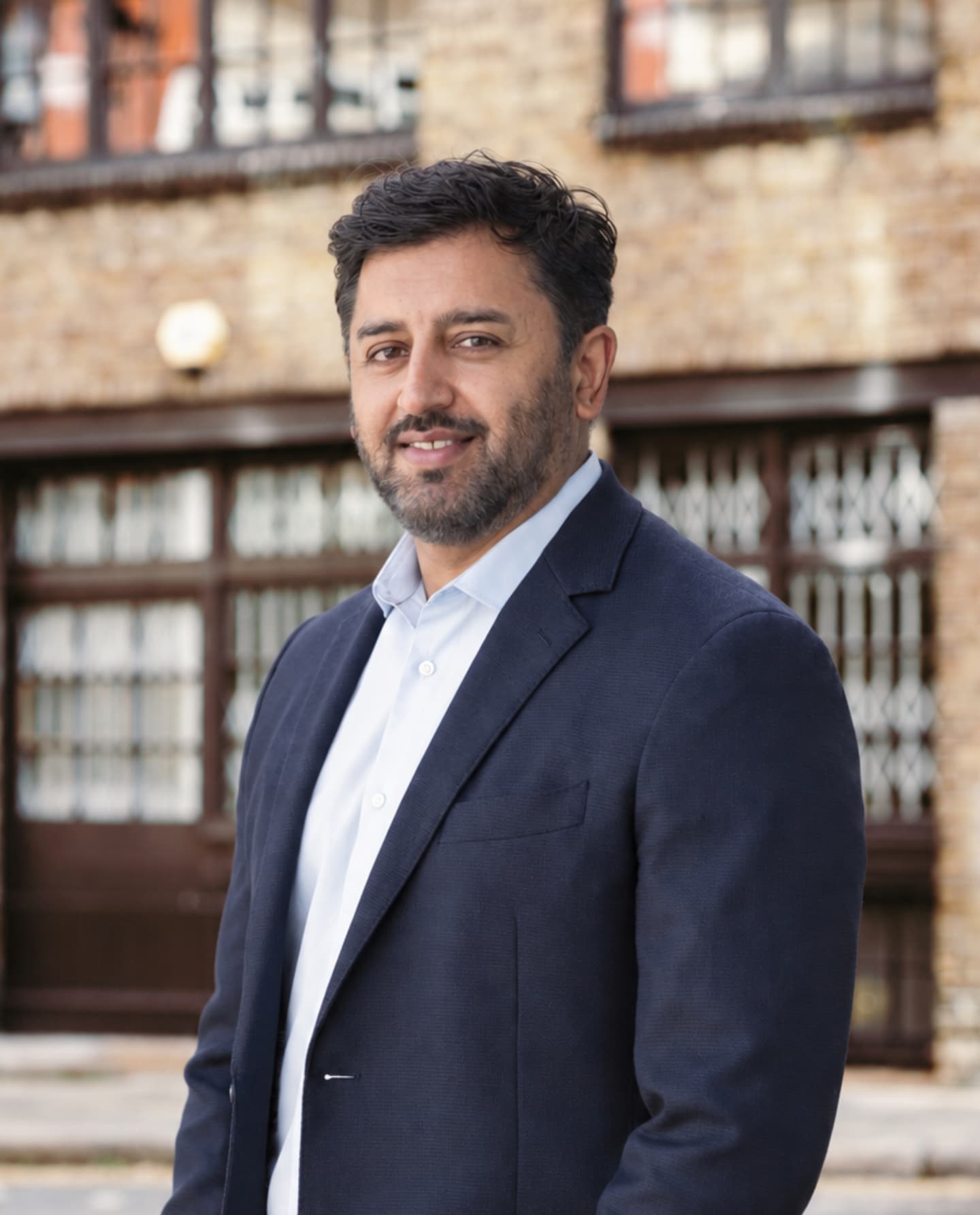 A smiling man with a beard, wearing a navy blazer and light blue button-down shirt.