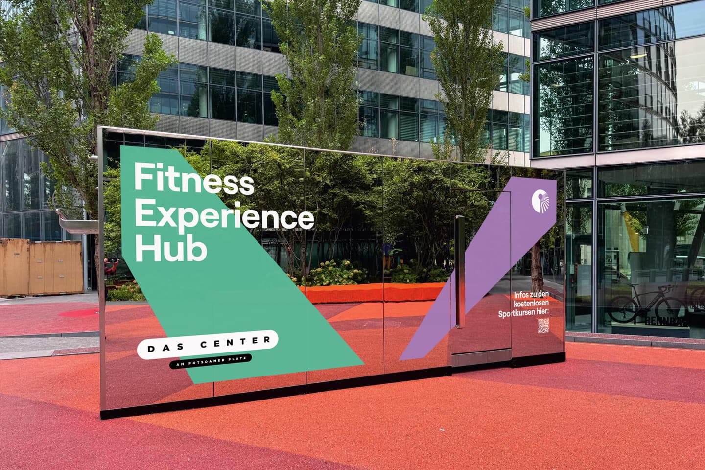 Outdoor glass structure labelled Fitness Experience Hub and DAS Centre stands on a red-paved area with trees and modern buildings in the background.