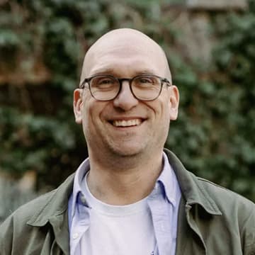 A man with glasses, a bald head, and a light-coloured shirt and jacket smiles outdoors with greenery in the background.