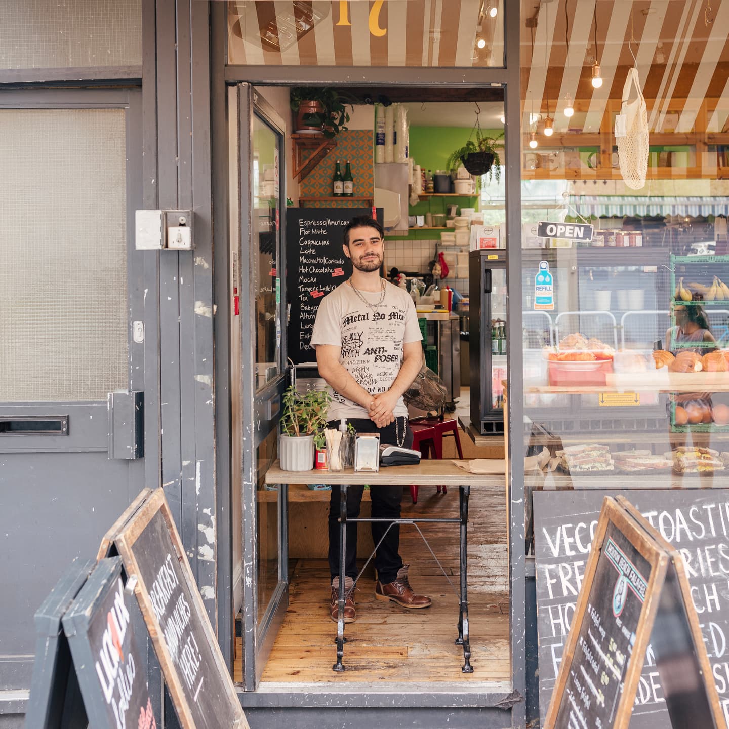 A man stands behind a small table at the entrance of a café, with chalkboard menus outside and shelves of food and plants visible inside.