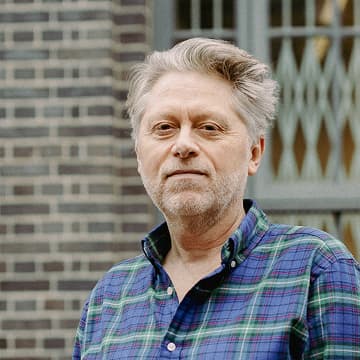 A middle-aged man with grey hair and a beard, wearing a green and blue checked shirt, stands in front of a brick building with a window in the background.